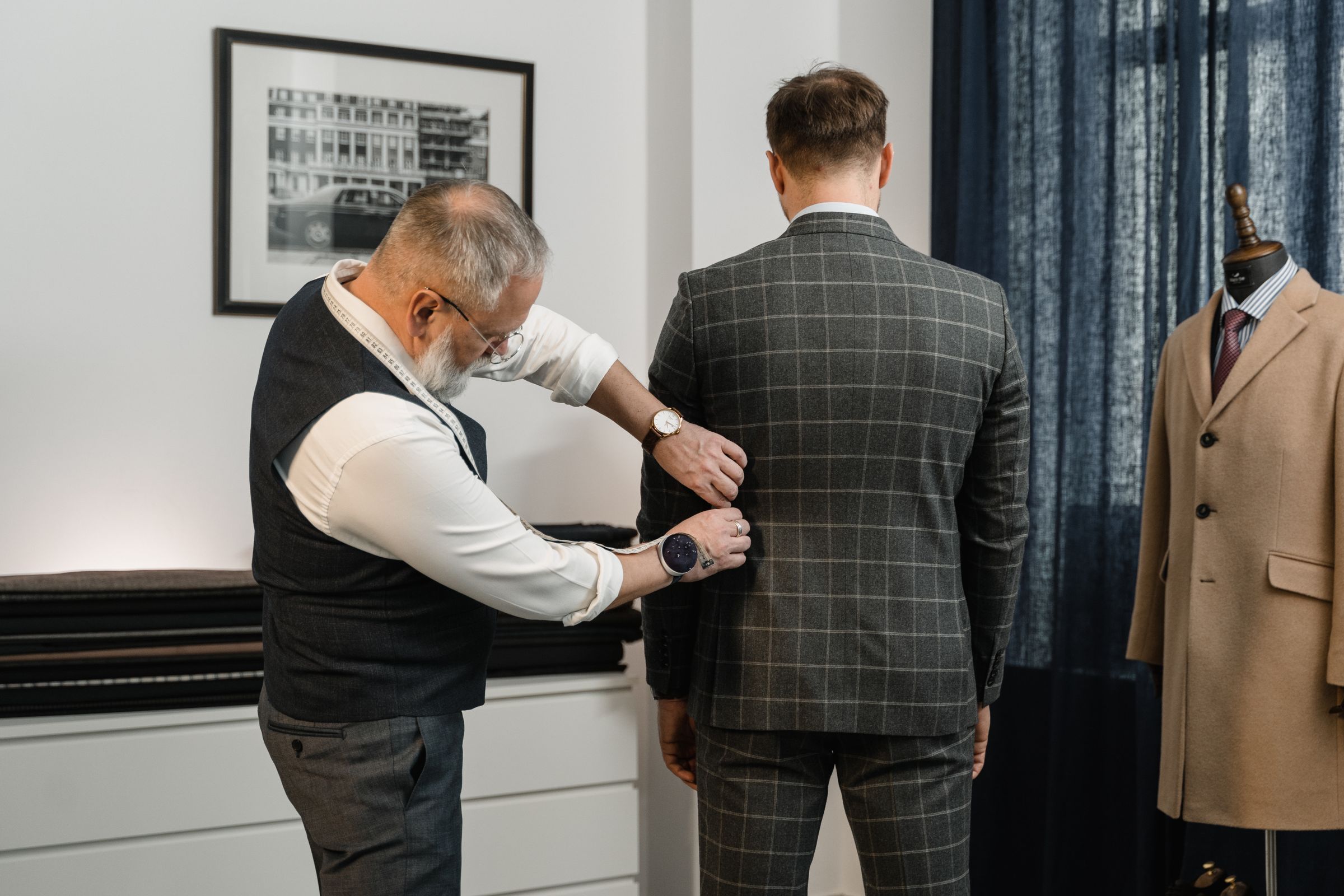 A tailor pins the back of a checked suit jacket during a fitting.